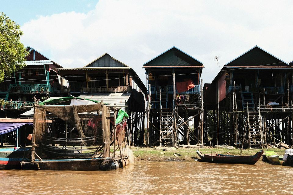 Una fila di case di legno su palafitte costeggia la riva di un fiume fangoso, con piccole barche e reti da pesca in primo piano.