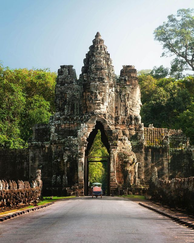 Un tuk-tuk percorre una strada attraverso un'antica porta di pietra con volti intagliati, affiancata da statue e circondata da una lussureggiante foresta verde.