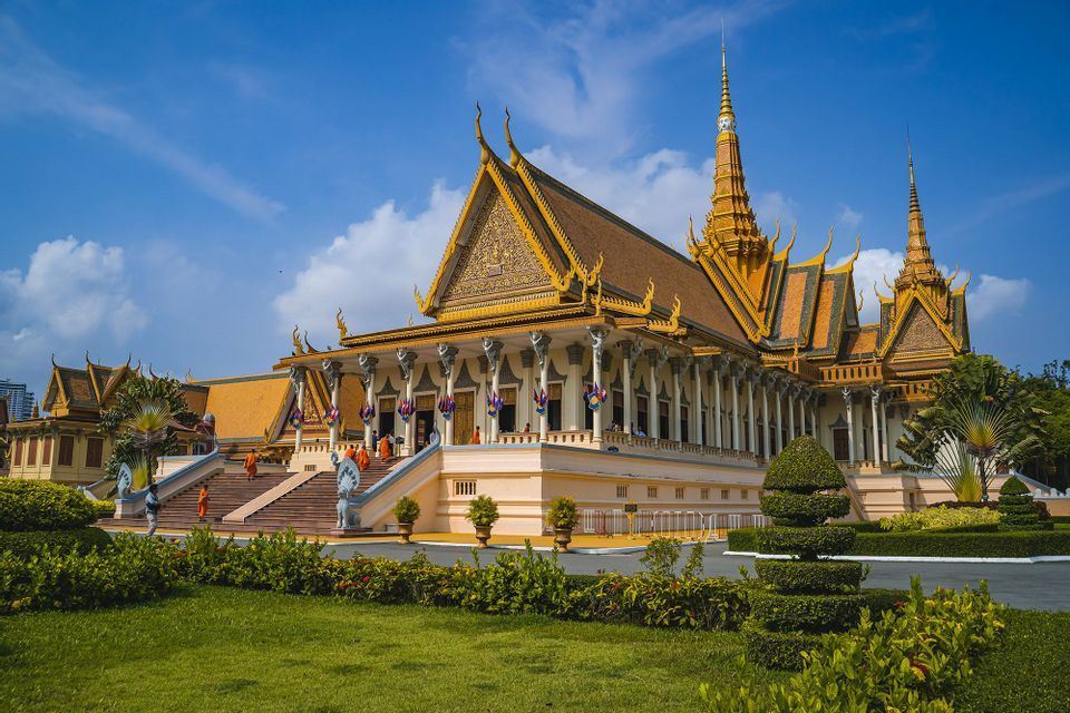 Diversi monaci in vesti arancioni salgono le scale di un tempio ornato con un tetto dorato a più livelli sotto un cielo blu.