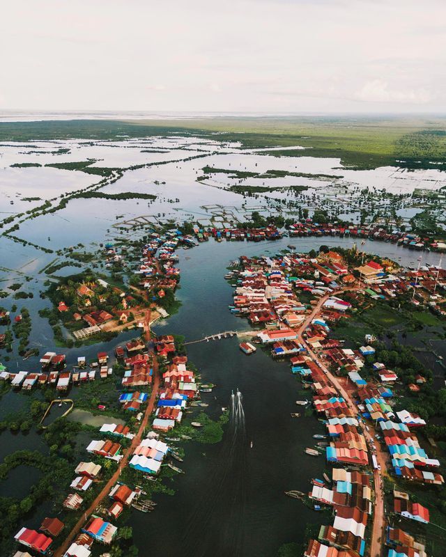 Una veduta aerea di un villaggio con case colorate su un fiume, circondato da una vasta pianura alluvionale e una foresta verde.