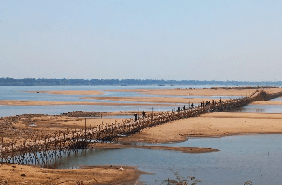Un gruppo WeRoad attraversa un lungo e rustico ponte di bambù su un ampio fiume sabbioso sotto un cielo limpido.