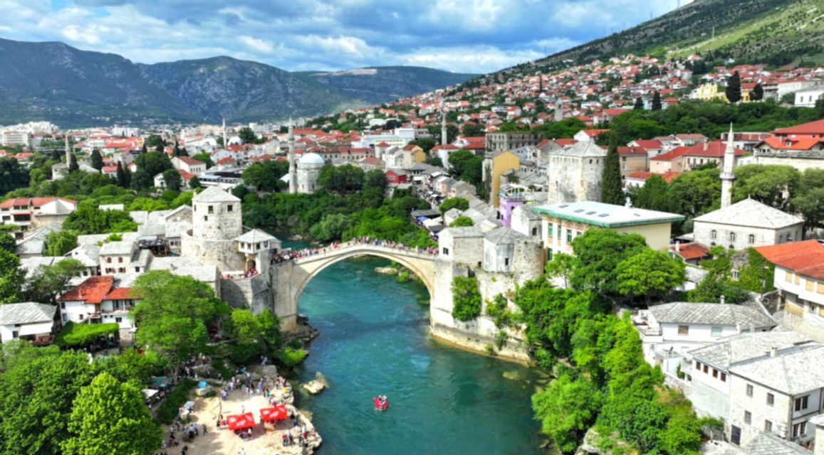 Luftbild einer historischen Stadt mit einer Steinbogenbrücke über einem türkisfarbenen Fluss, mit Häusern und Bergen im Hintergrund.