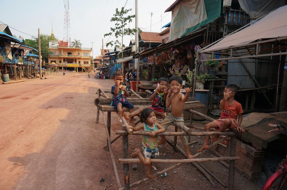 Un gruppo di bambini è seduto su una struttura di legno rustica, lungo una strada sterrata in un villaggio, sorridendo e mettendosi in posa per la fotocamera.