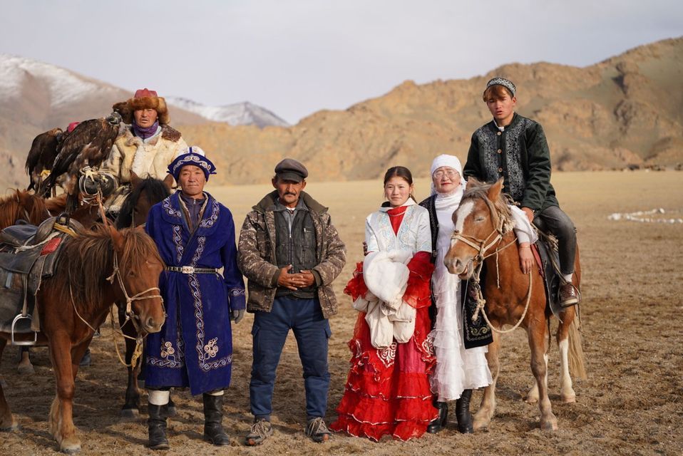 Un gruppo di persone in abiti tradizionali posa per una foto con cavalli e un'aquila reale in un vasto paesaggio collinare.
