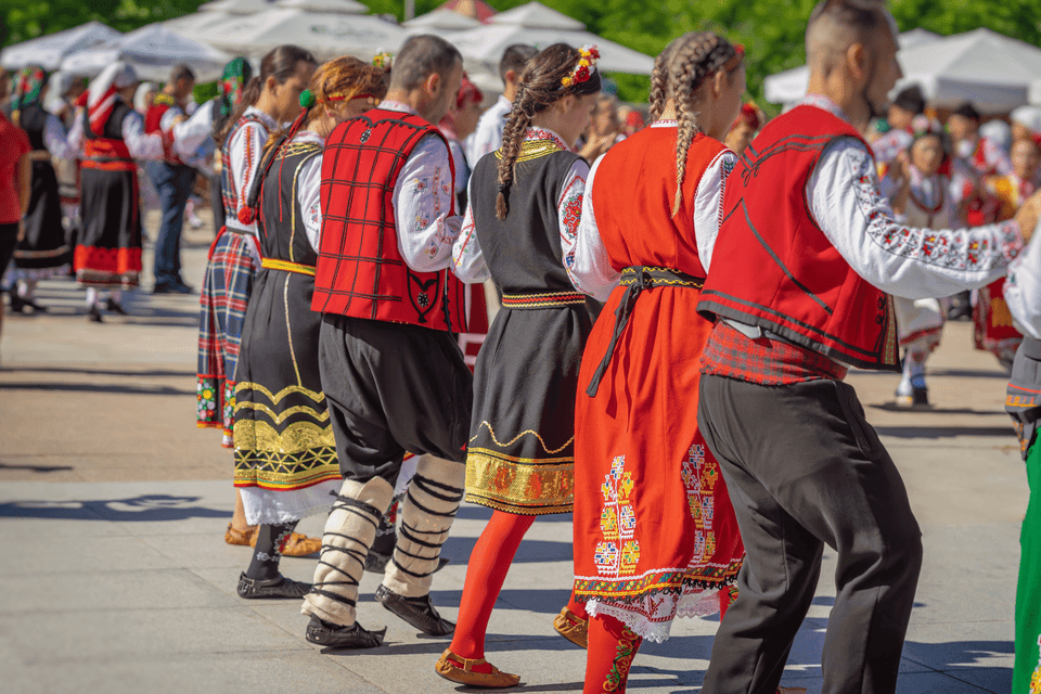 Una fila de bailarines folclóricos con coloridos trajes tradicionales bordados actúan al aire libre.