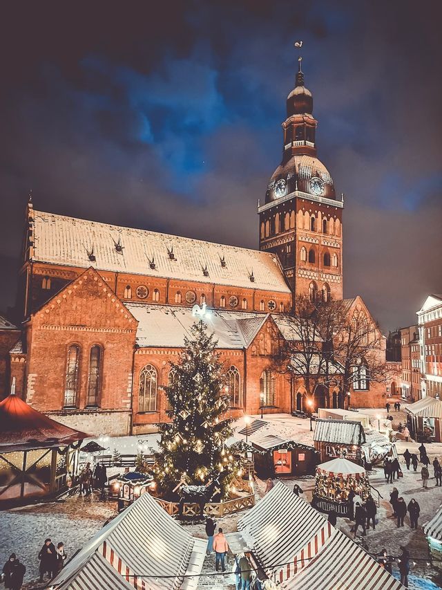 Une place de ville enneigée la nuit accueille un marché de Noël avec un grand sapin décoré, devant une cathédrale en briques illuminée.