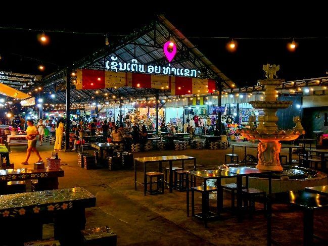 Un marché nocturne en plein air avec une fontaine décorative au premier plan, rempli de tables et d'étals sous des guirlandes de lumières chaudes.
