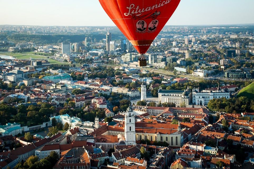 Un globo aerostático rojo con gente en su cesta flota sobre un vasto paisaje urbano con edificios de tejados rojos y rascacielos modernos.