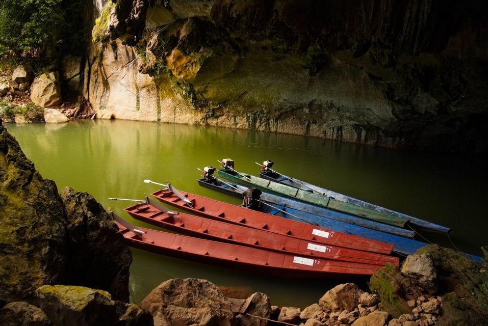 Plusieurs bateaux à longue queue rouges, bleus et verts sont amarrés sur une eau vert trouble, à l'intérieur d'une grotte rocheuse.