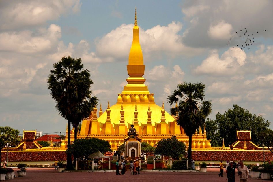 Un grand stupa doré avec une haute flèche centrale se dresse dans une cour, flanqué de palmiers sous un ciel partiellement nuageux.