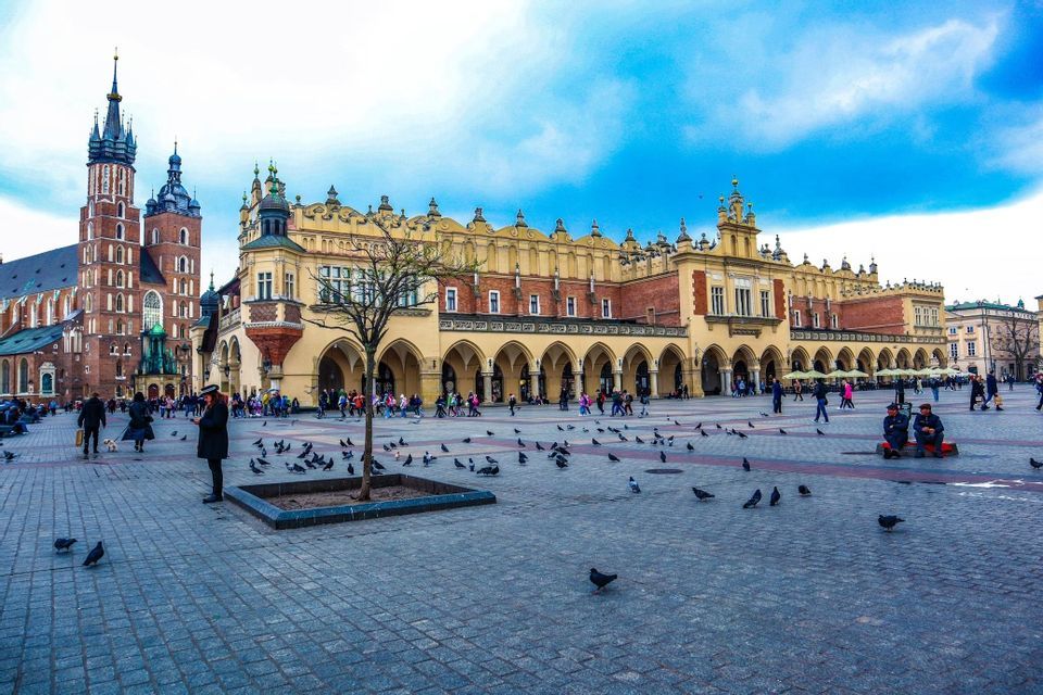 Una animada plaza histórica de la ciudad con gente y palomas en una plaza adoquinada frente a edificios antiguos y ornamentados.