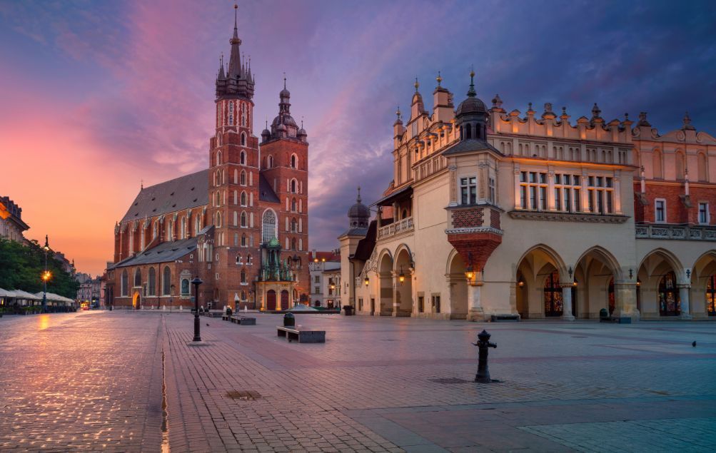 Una plaza de ciudad adoquinada vacía con una alta iglesia de ladrillo y un salón ornamentado bajo un cielo de amanecer colorido.