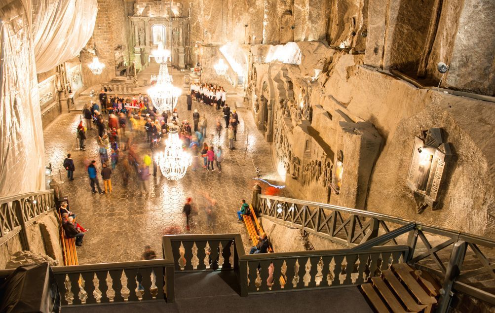 Vista aérea y de larga exposición de personas en una vasta cámara subterránea excavada en roca, iluminada por grandes candelabros.