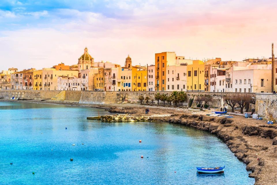 Edifici colorati di una cittadina costiera si allineano lungo un molo in pietra, accanto a una tranquilla baia blu con una piccola barca.