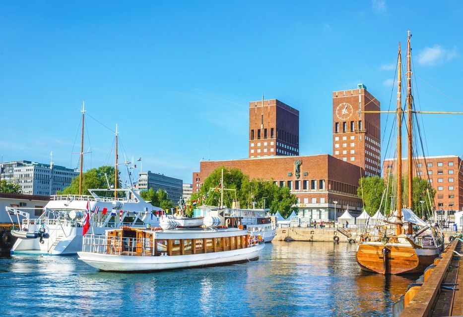 Diverse barche e yacht sono ormeggiati in un porto cittadino, di fronte a un grande edificio in mattoni rossi con due torri, sotto un cielo azzurro.