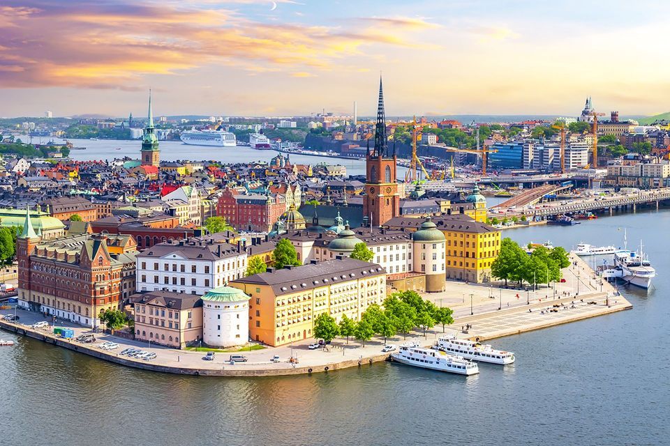 Veduta aerea di una storica città europea sull'acqua, con edifici colorati, una guglia di chiesa e barche ormeggiate lungo la riva al tramonto.