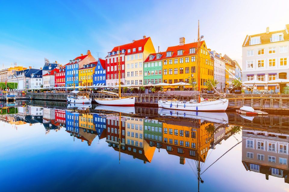 Edifici colorati e barche ormeggiate lungo un porto, perfettamente riflesse nell'acqua calma sotto un cielo azzurro e limpido.