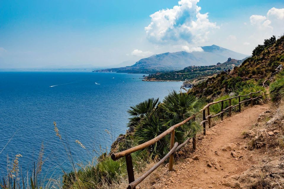 Un chemin de terre avec une clôture en bois longe une falaise côtière surplombant la mer bleue et une chaîne de montagnes lointaine.