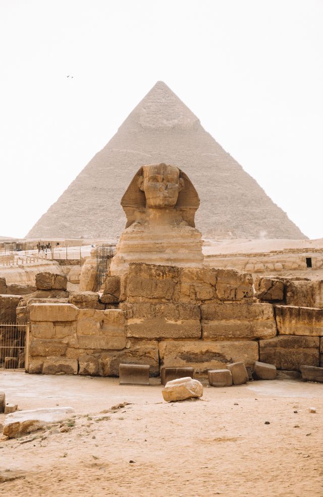 Le Grand Sphinx se dresse devant une grande pyramide dans un paysage sablonneux, avec des ruines de pierre antiques au premier plan.