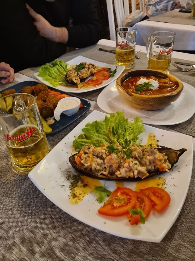 Ein Tisch gedeckt mit verschiedenen Gerichten, darunter gefüllte Auberginen und Eintopf, zusammen mit mehreren Gläsern Bier.
