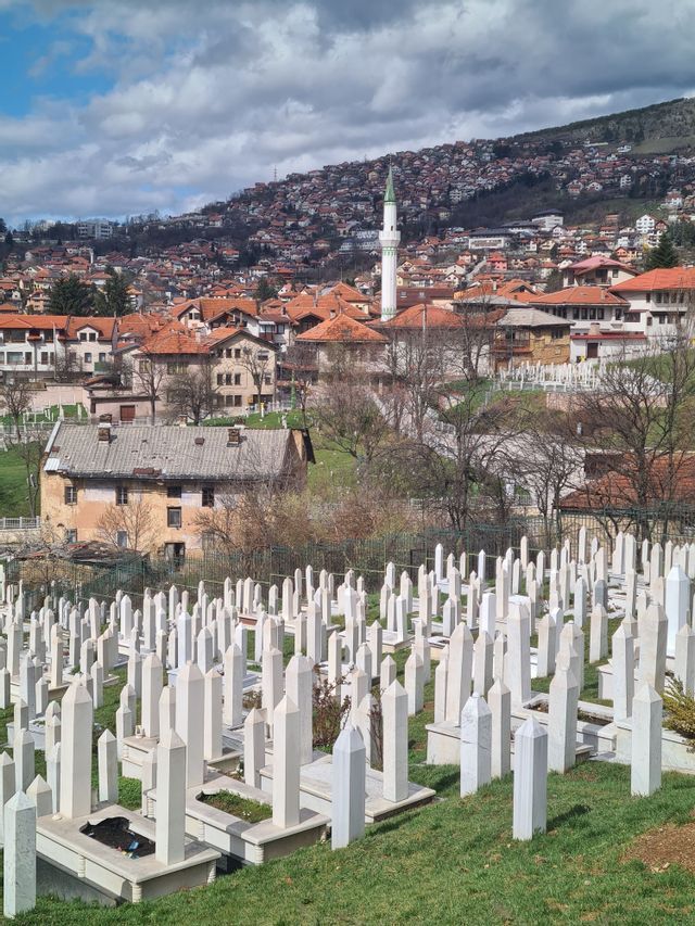Ein Friedhof am Hang mit weißen Grabsteinen überblickt eine Stadt mit roten Dächern und einem Minarett unter einem bewölkten Himmel.