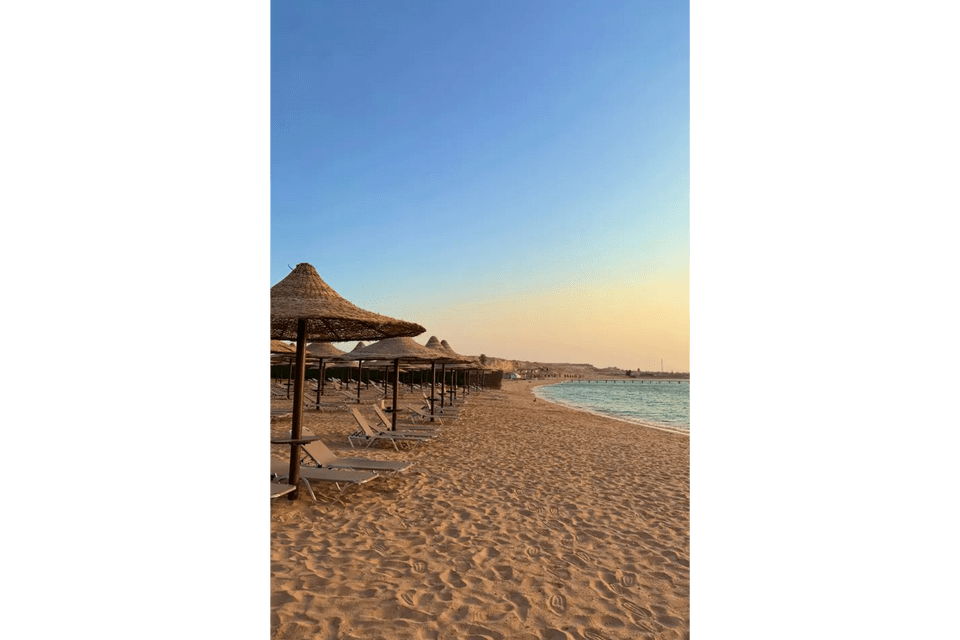 Una fila di lettini vuoti e ombrelloni di paglia fiancheggia una spiaggia sabbiosa in riva al mare al tramonto.