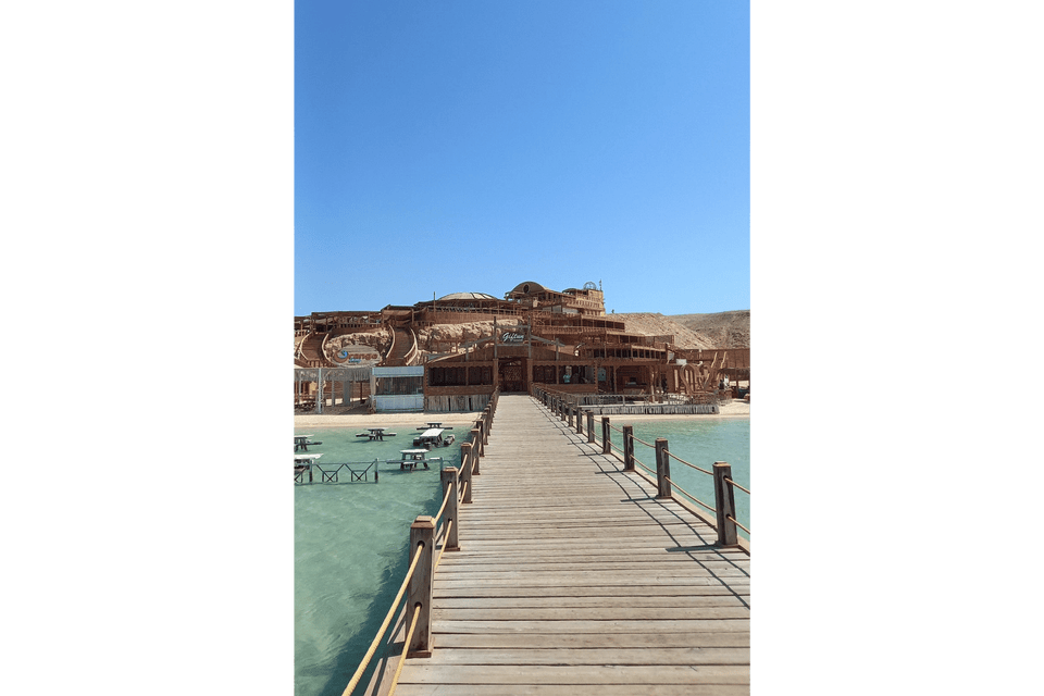 Un lungo molo di legno si estende su acque turchesi e limpide verso un grande resort balneare in legno costruito su un versante sabbioso sotto un cielo azzurro e sereno.