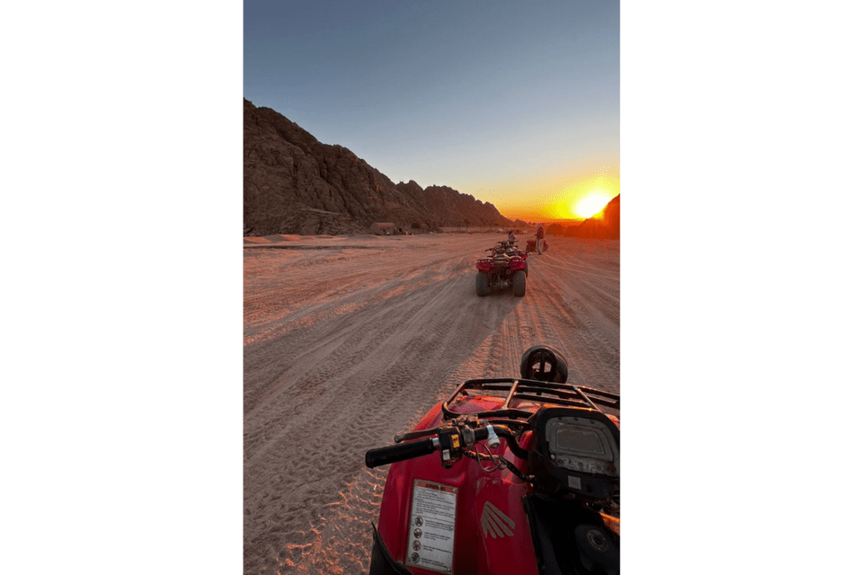 Vista in prima persona da un quad rosso durante un viaggio di gruppo WeRoad su una pista sabbiosa nel deserto al tramonto, con montagne sullo sfondo.