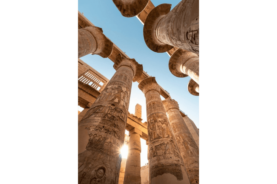 Vista dal basso di imponenti colonne di pietra con geroglifici egizi, con il sole che splende in un cielo azzurro e limpido.