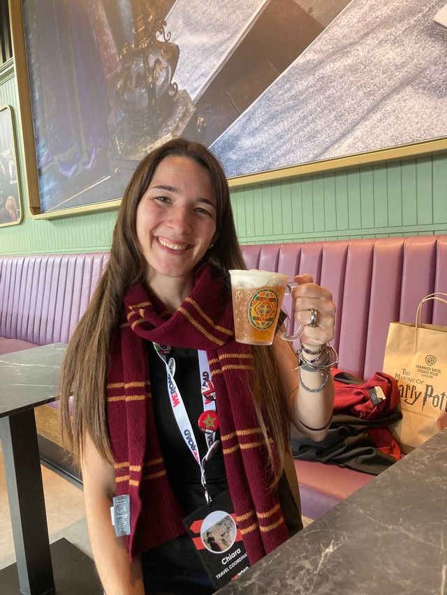 Una donna sorridente con una sciarpa a righe rosse e oro e un laccetto WeRoad solleva un boccale di vetro con una bevanda schiumosa.