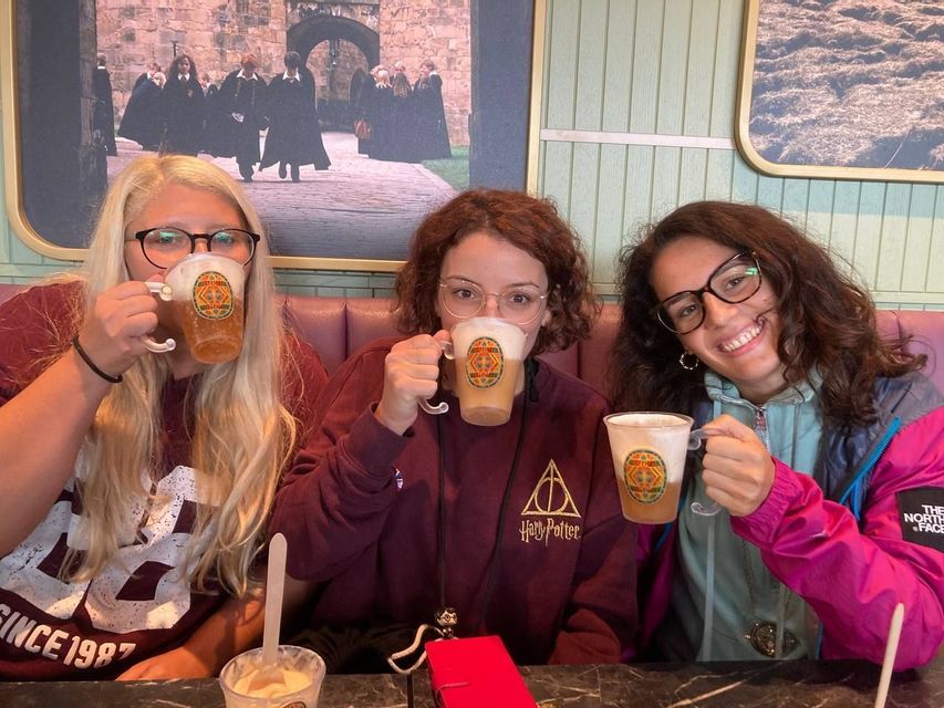 Tre donne di un gruppo WeRoad bevono da tazze in un caffè a tema, una con una felpa di Harry Potter.