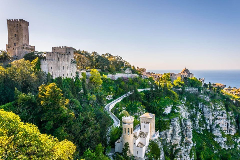 A stone castle sits atop a lush, green cliffside overlooking the ocean on a sunny day.