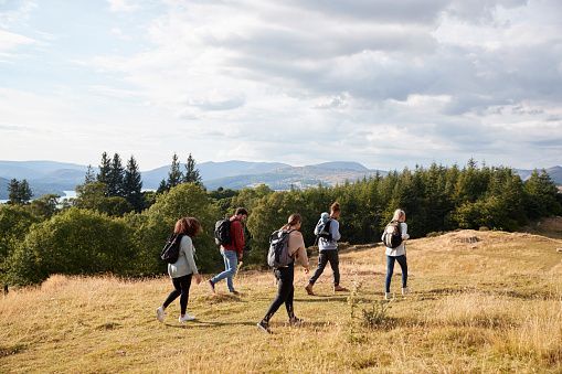 A WeRoad group trip of people with backpacks hiking on a grassy hill with a view of a forest, a lake, and mountains.
