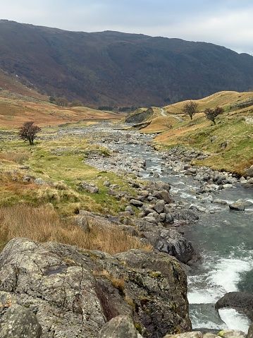 A rocky river flows through a grassy valley at the foot of a large mountain under an overcast sky.