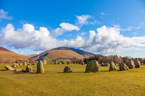A circle of large standing stones sits in a grassy field with rolling hills and mountains in the background under a blue sky with white clouds.