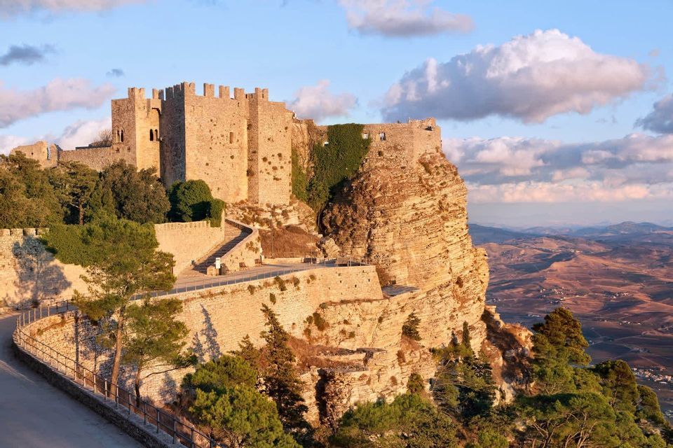 Un château de pierre perché sur une falaise rocheuse surplombant une vallée, illuminé par la lumière chaude du coucher de soleil.