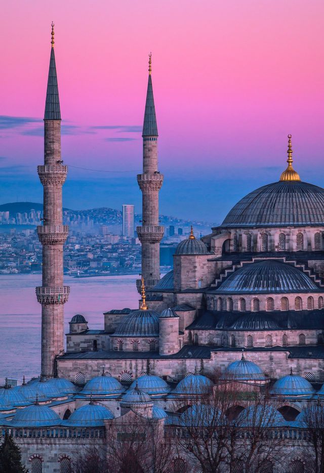 A large mosque with multiple blue domes and two tall minarets overlooks a city and water under a pink and purple sky at dusk.