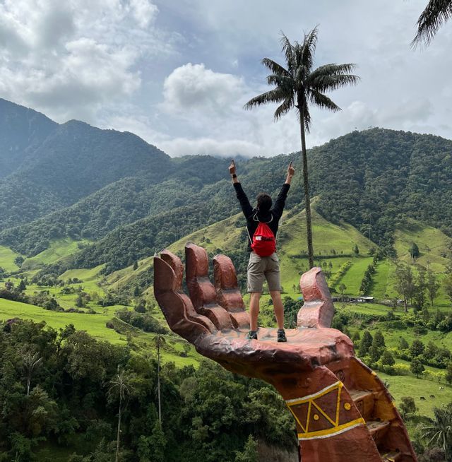 Eine Person mit rotem Rucksack steht auf einer großen Handskulptur mit erhobenen Armen, mit Blick auf ein üppiges grünes Tal und eine hohe Palme.