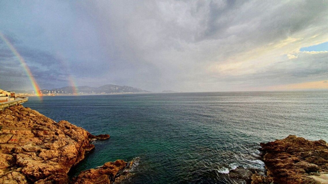Un double arc-en-ciel s'étend au-dessus de la silhouette d'une ville lointaine, de l'autre côté de la mer, vue depuis un littoral rocheux sous un ciel nuageux.