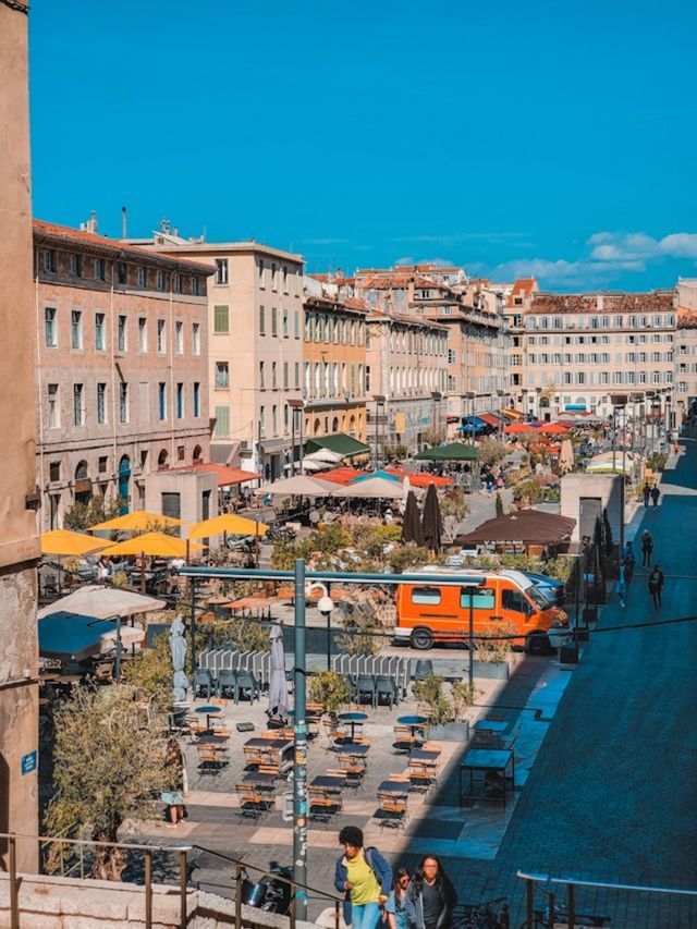 Une vue plongeante sur une place de ville animée avec des cafés en plein air, des parasols colorés et des bâtiments historiques sous un ciel bleu clair.