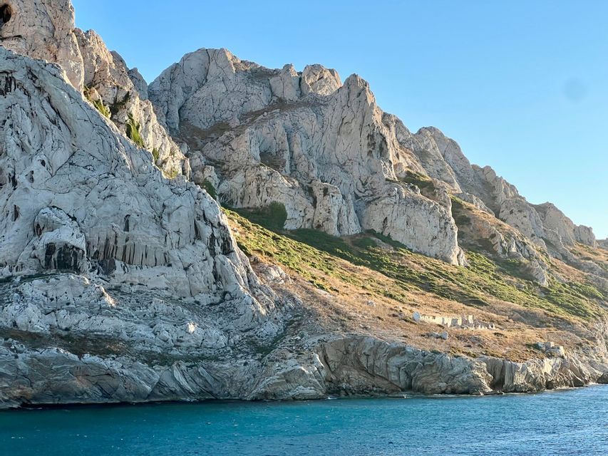 Une chaîne de montagnes accidentées et rocheuses, avec des pentes herbeuses, descend vers la mer calme et bleue sous un ciel clair.