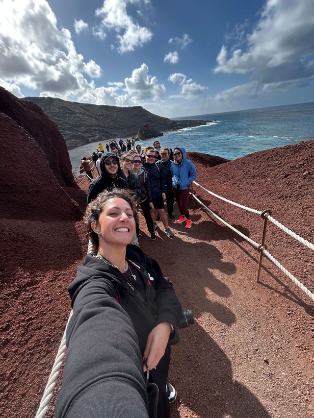 Una mujer se hace un selfie con su viaje en grupo de WeRoad en un sendero costero con tierra roja junto al mar azul.
