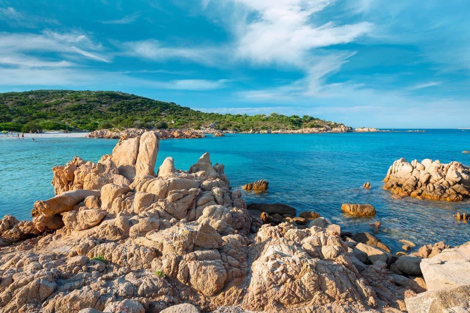 Una costa rocciosa si affaccia su una baia con acque turchesi e una lontana spiaggia di sabbia bianca sotto un cielo azzurro parzialmente nuvoloso.