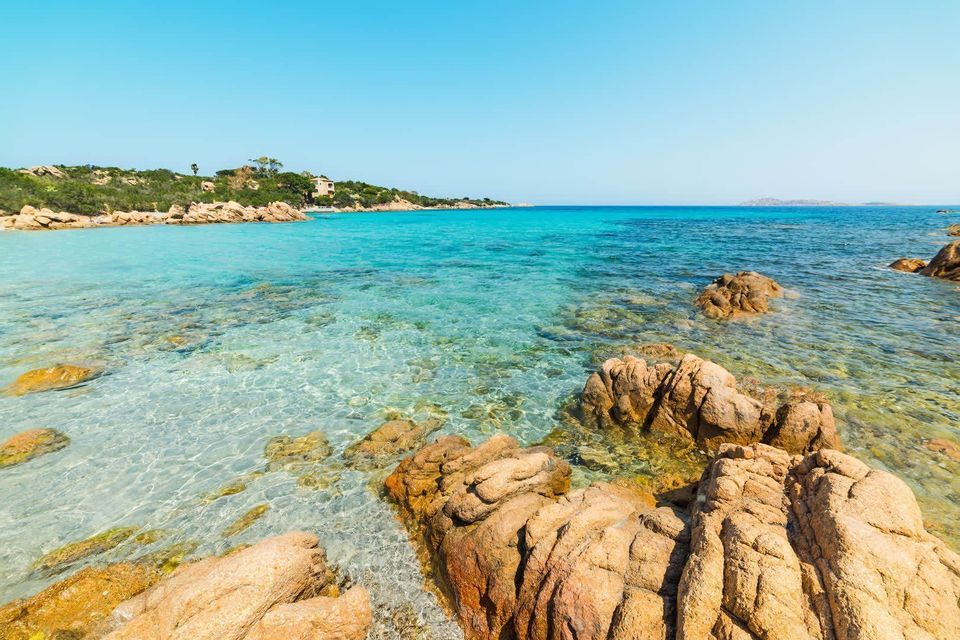 Clear, turquoise sea water washing over large brown rocks on a sunny coastline under a bright blue sky.