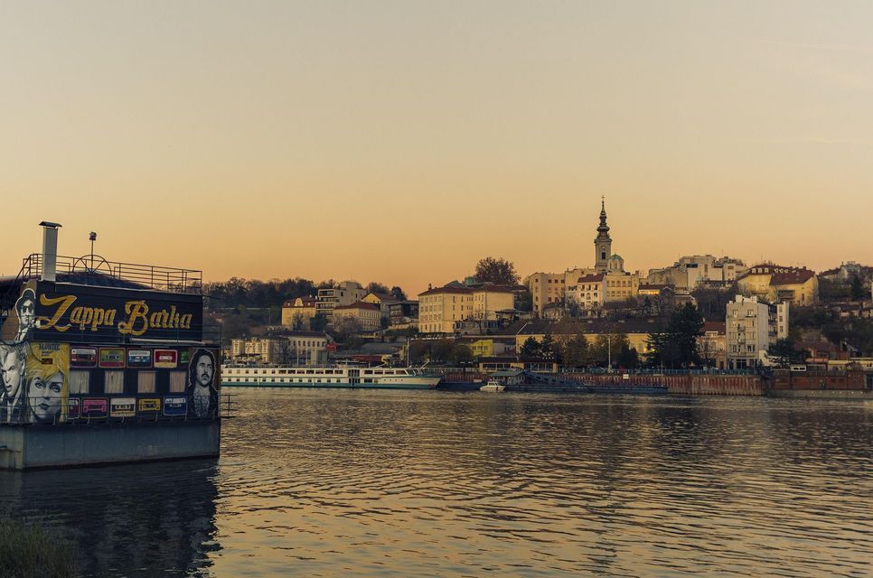 Una casa flotante llamada Zappa Barka flota en un río con el horizonte de una ciudad, incluyendo un campanario de iglesia, visible al fondo al atardecer.