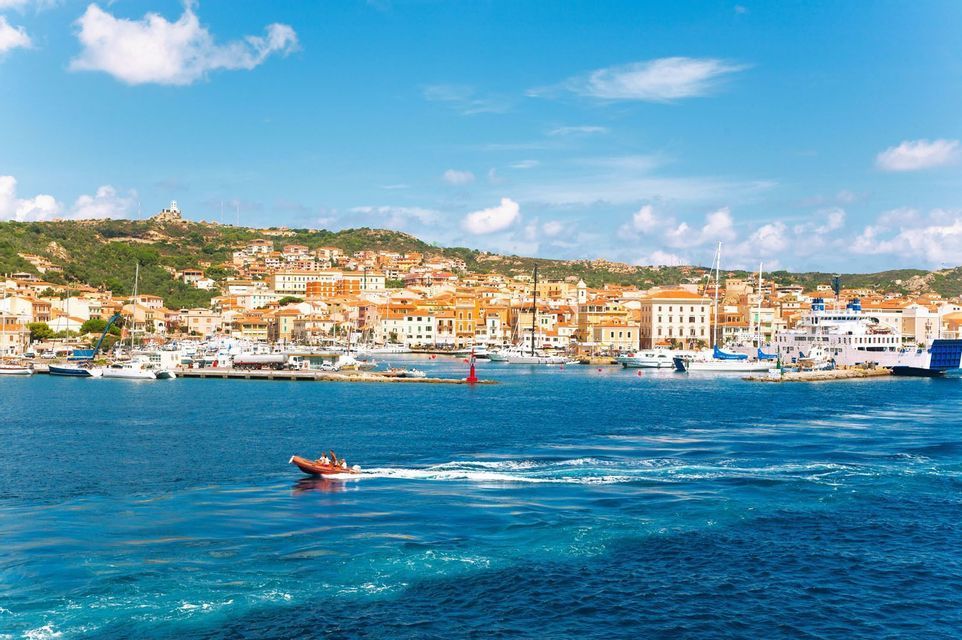 A small motorboat travels across blue water in front of a coastal town's harbor, which is filled with docked boats under a partly cloudy sky.