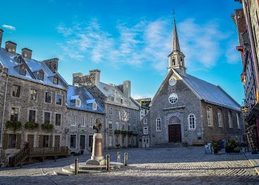 Une église historique en pierre avec un clocher se dresse sur une place pavée, entourée de bâtiments en pierre à plusieurs étages sous un ciel bleu éclatant.