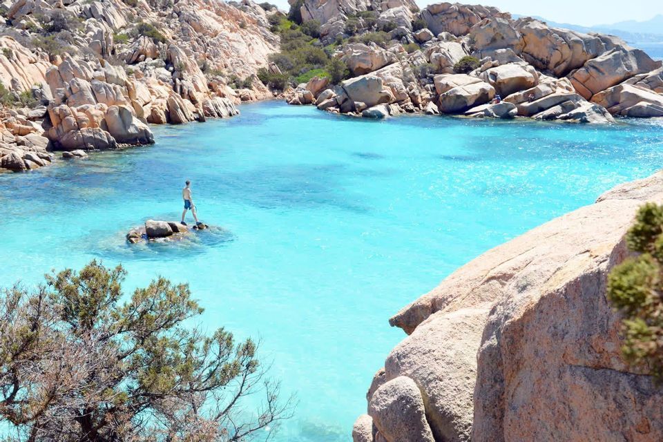 A man stands on a rock in the middle of a cove with turquoise water, surrounded by rocky cliffs.