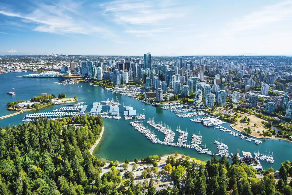 Una vista aérea de un horizonte urbano moderno que bordea una bahía llena de marinas y barcos atracados, con un denso bosque verde en primer plano.