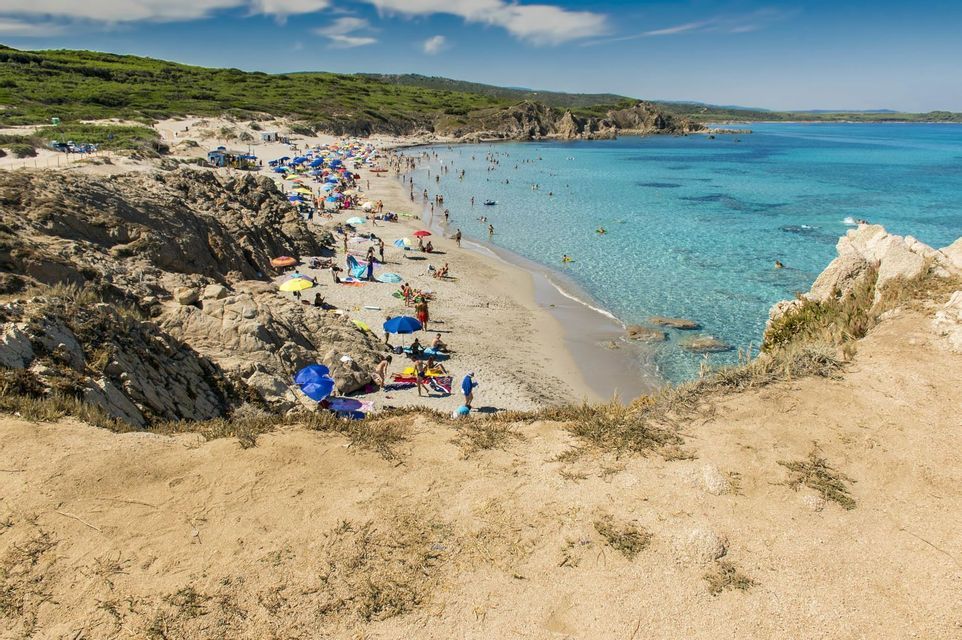 Vue plongeante sur une plage de sable bondée, avec des gens qui nagent dans une eau turquoise cristalline et se détendent sous des parasols colorés.